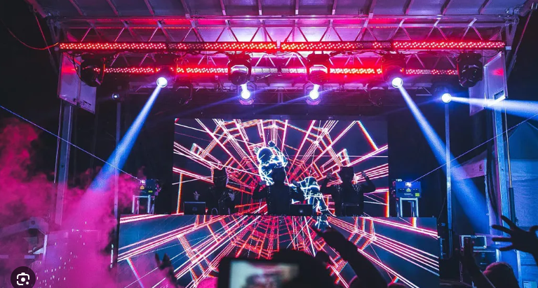 Escenario con DJ, luces brillantes y efectos visuales geométricos, creando una atmósfera de concierto con niebla.