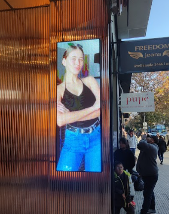 Pantalla digital con una mujer vestida con top negro y jeans, frente a una tienda con el letrero 