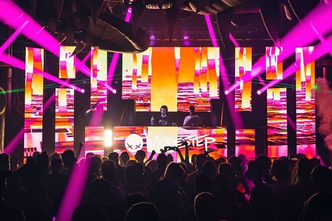 Un DJ actuando en una discoteca con público, iluminada con luces rosas y naranjas.