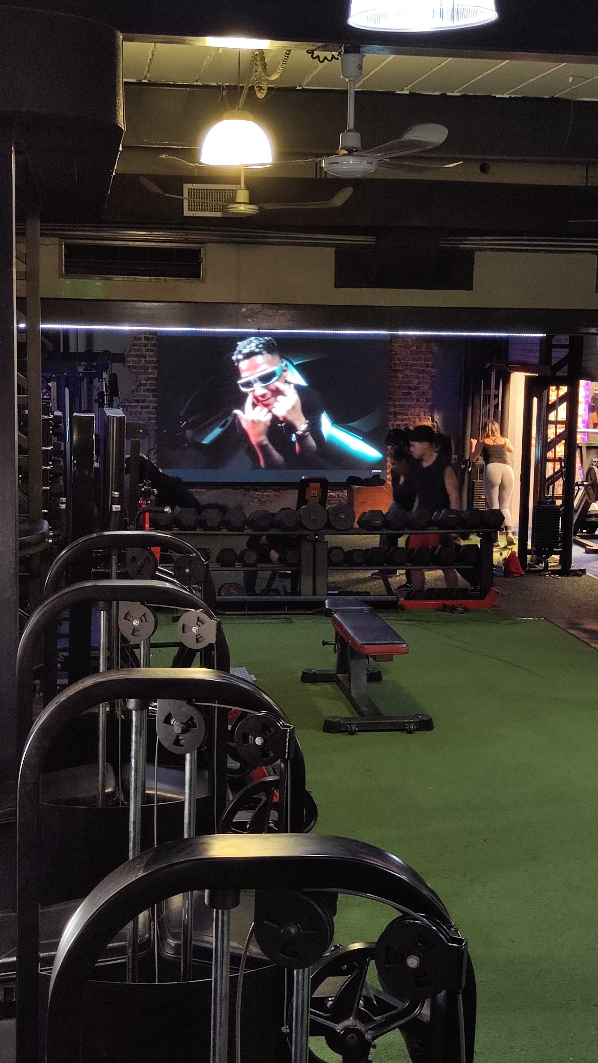 Interior del gimnasio con máquinas de pesas, bancos y una pantalla grande que reproduce un vídeo.