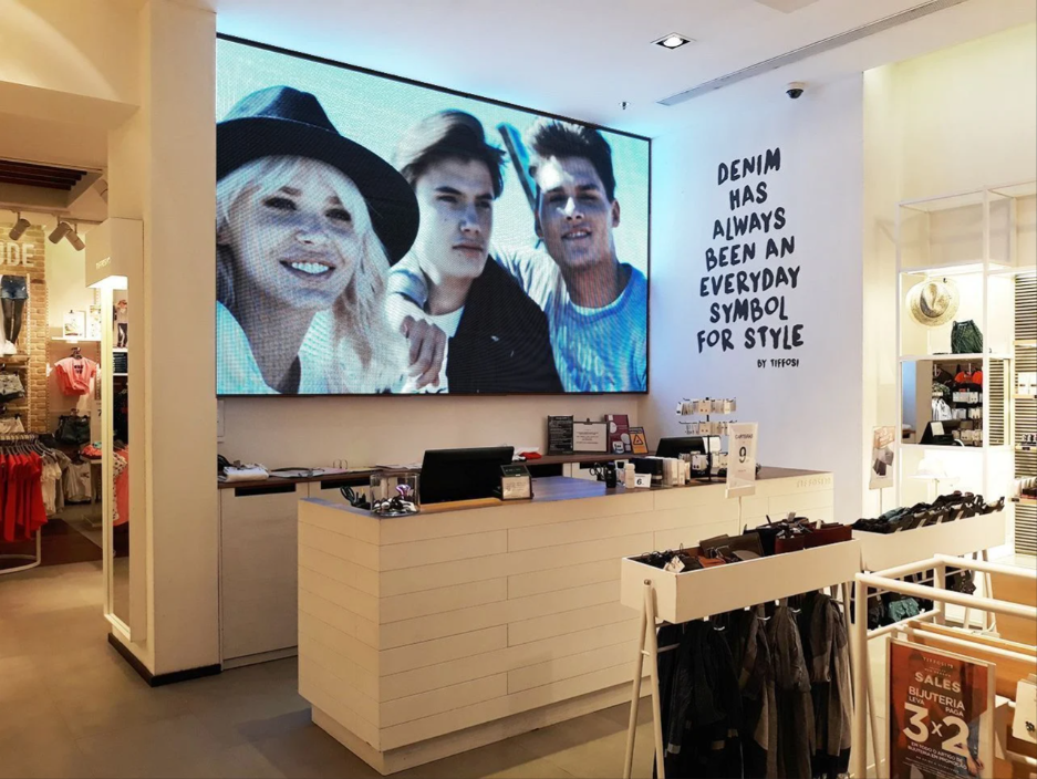Interior de una tienda de ropa con una gran pantalla que muestra a tres jóvenes. Se ven la caja registradora y los percheros.