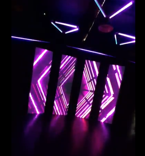 Luces de neón púrpura iluminan una habitación oscura; formas geométricas decoran las paredes y el techo.