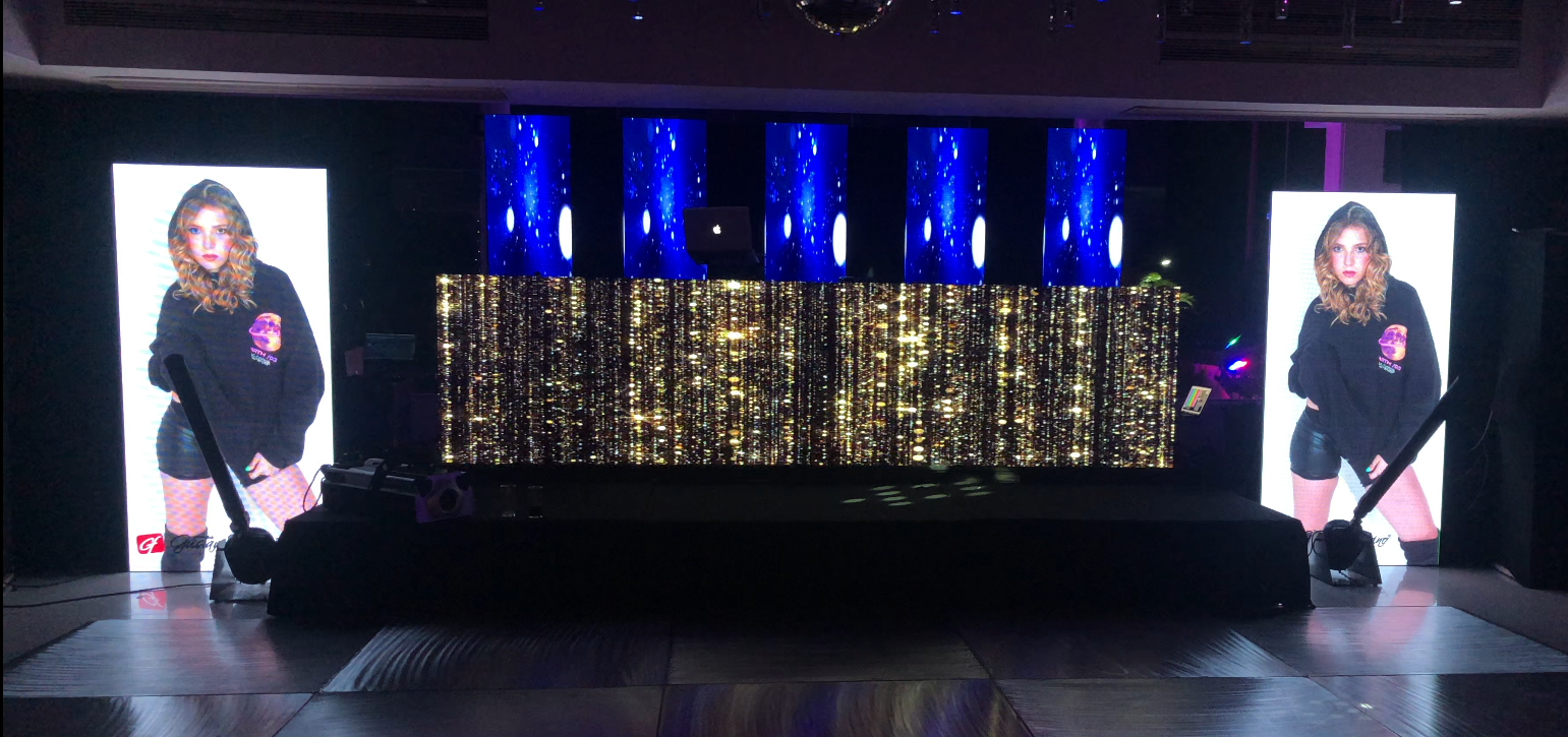 Escenografía con dos pantallas que muestran a una persona, luces azules y flecos dorados.