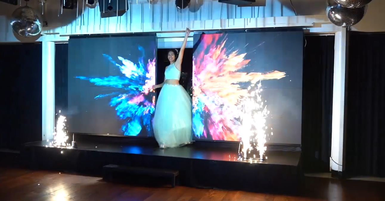 Mujer con vestido azul verdoso en el escenario con un colorido espectáculo de luces y bengalas.