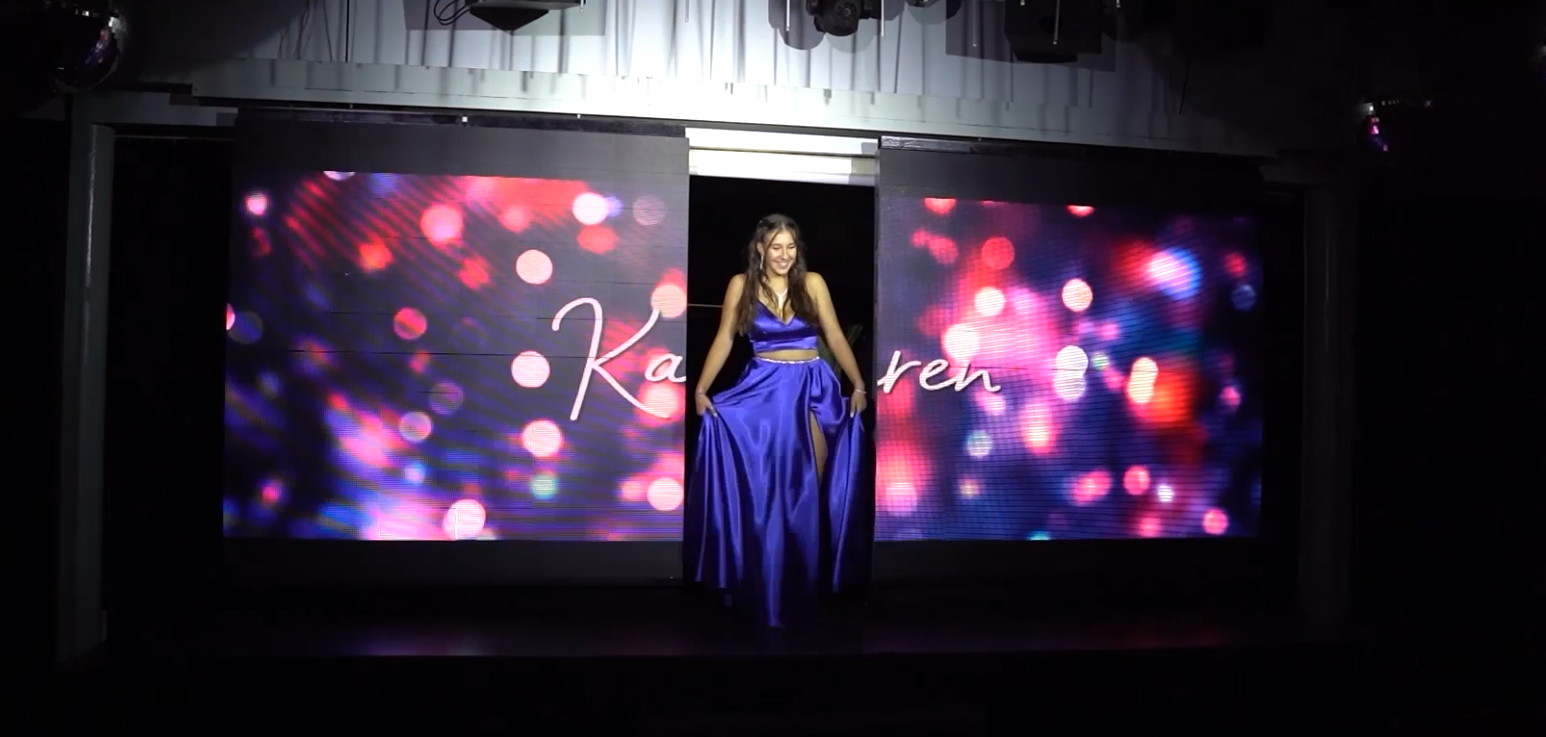 Una mujer con vestido azul está de pie en el escenario con un telón de fondo que muestra el texto 