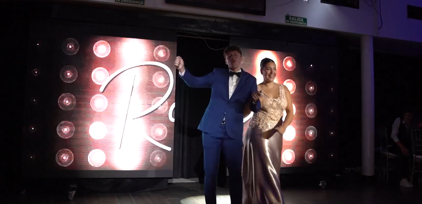 Un hombre con traje azul y una mujer con vestido dorado están de pie frente a una pantalla iluminada.