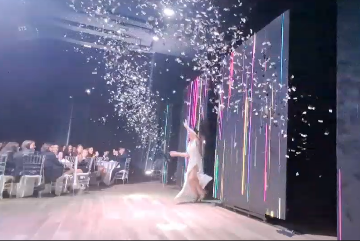 Mujer vestida de blanco en el escenario con confeti. Personas sentadas a mesas al fondo. Espectáculo de luces de colores.