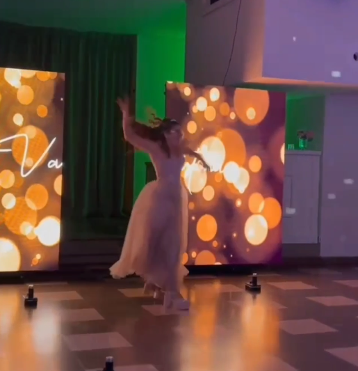 Una mujer vestida de blanco baila en una pista de baile iluminada con luces y pantallas doradas.