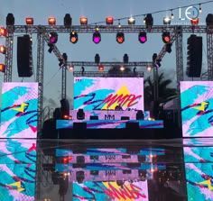 Escenario montado con luces, pantallas y equipo de DJ; colores vivos, entorno al aire libre.