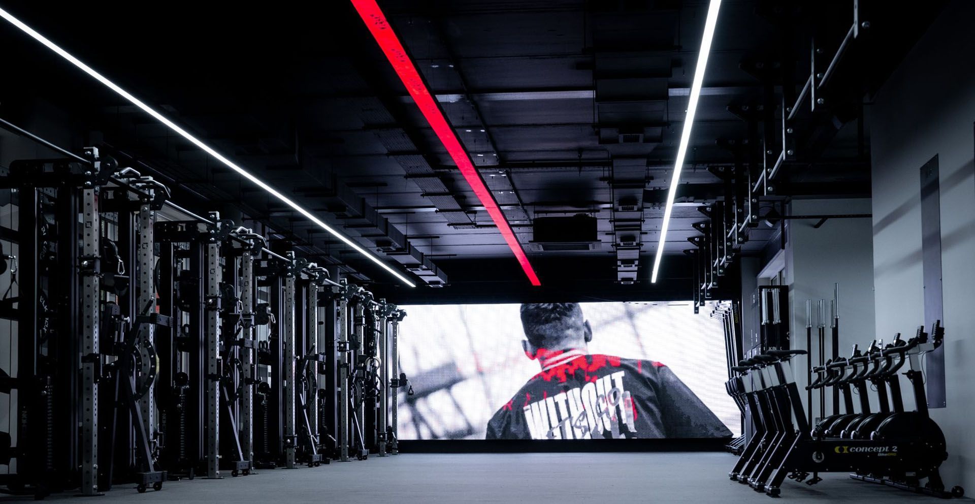 Gimnasio con equipamiento, una gran pantalla que muestra a una persona, iluminación superior roja y blanca.