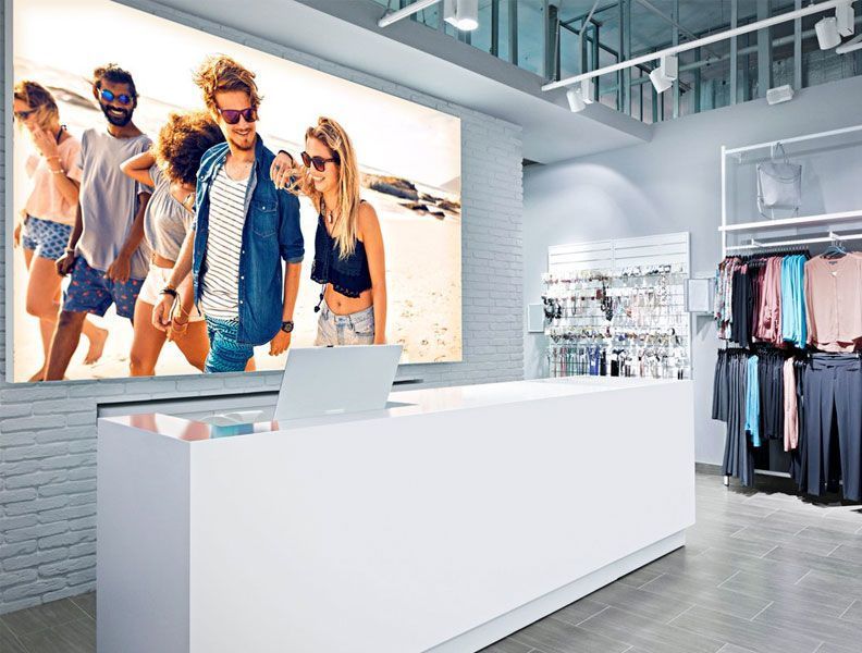 Interior de una tienda de ropa con mostrador blanco, percheros y fotografía retroiluminada de personas en la playa.