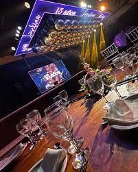 Una mesa de banquete puesta para una celebración. Copas de champán, platos y centros de mesa con un escenario al fondo.