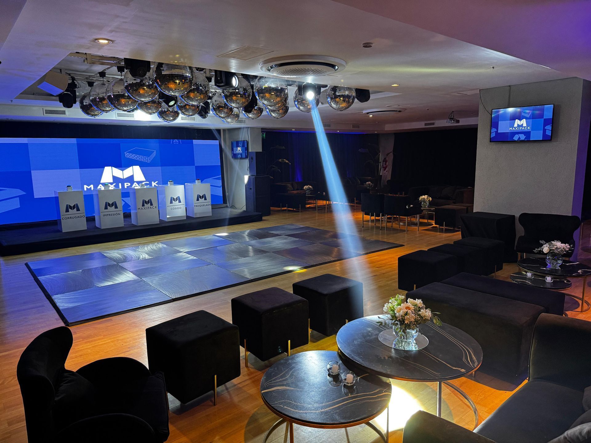 Espacio para eventos: escenario con logotipo, bolas de discoteca, asientos y mesas. 