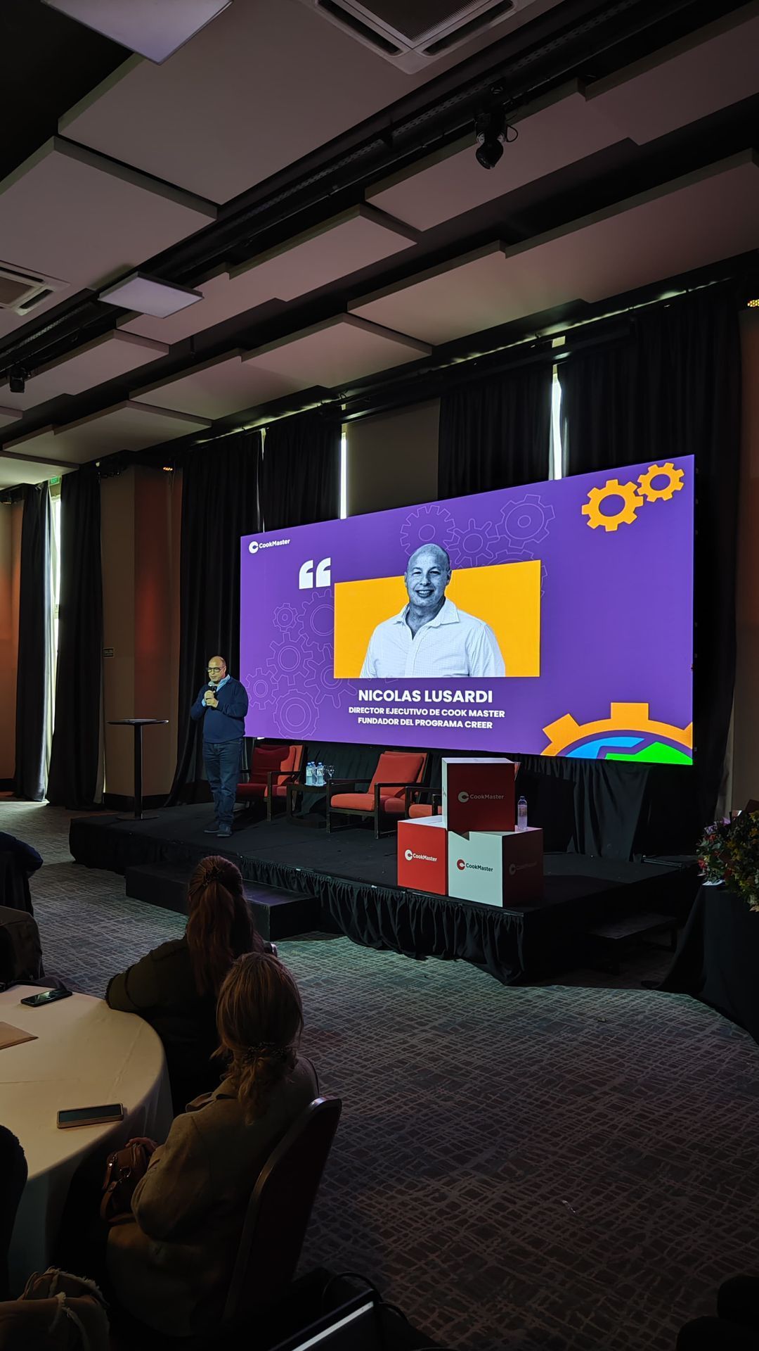 Hombre hablando en el escenario con una pantalla que muestra la imagen del orador y una cita, público sentado.