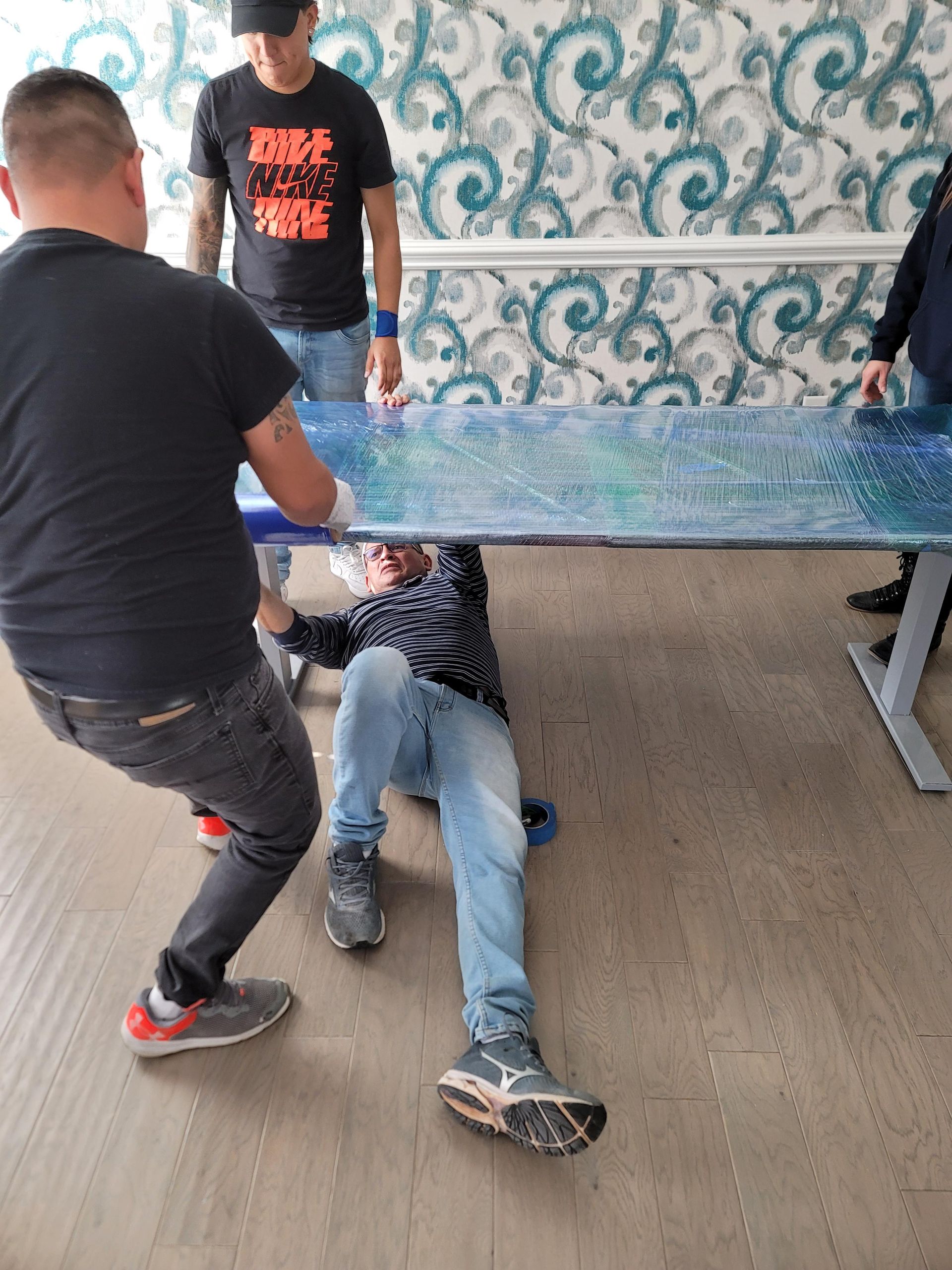 A man is laying on the floor under a table being lifted by two men.