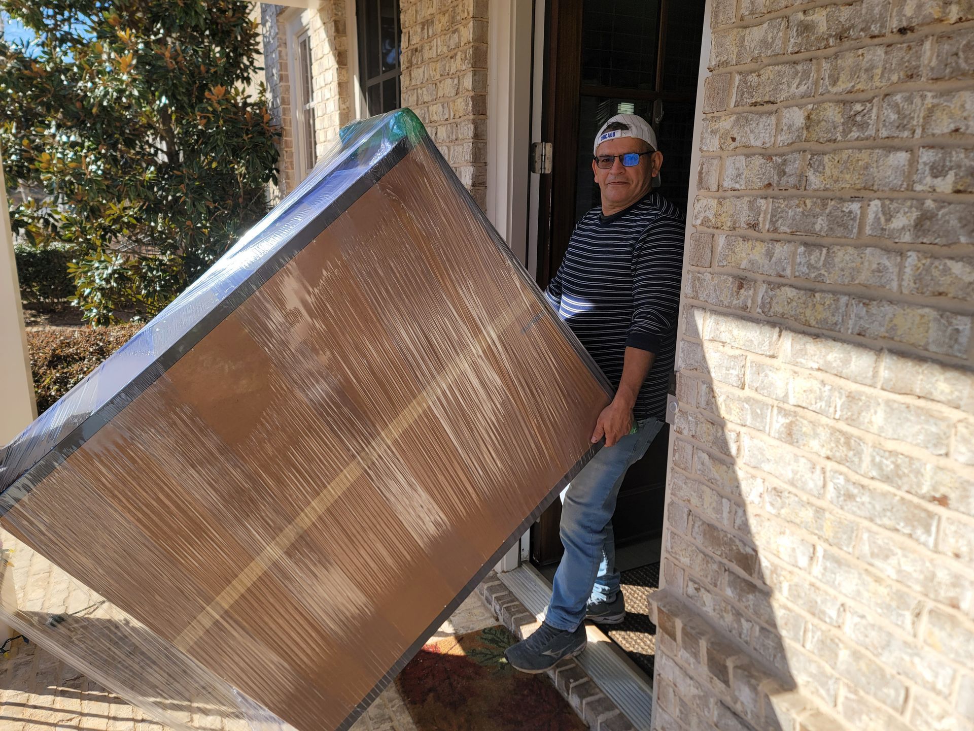 A man is carrying a large piece of wood in front of a brick building.