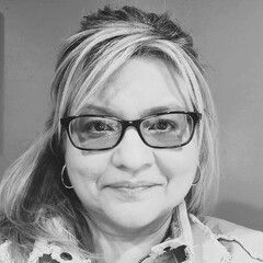 Woman with blonde and gray hair, wearing glasses, smiling, hoop earrings, in a well-lit room.