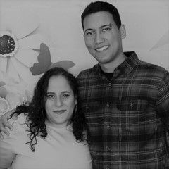 Two people smiling at the camera. Man in plaid shirt, woman in light shirt, floral backdrop.