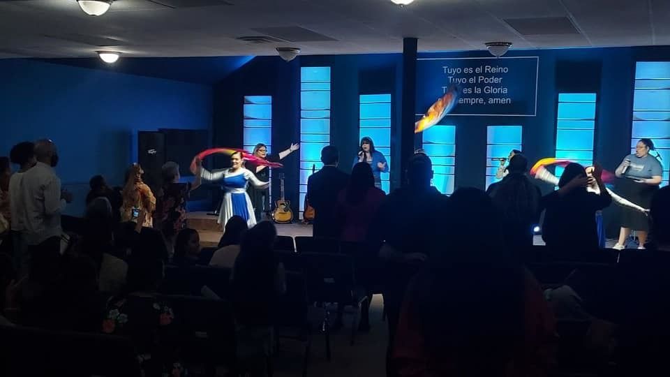 People worshiping in a church with a band on stage; blue lighting, stage has text.