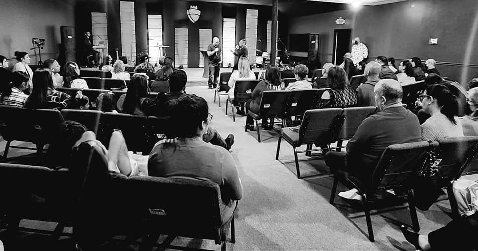 Audience in a church service, listening to speakers on a stage. Black and white.