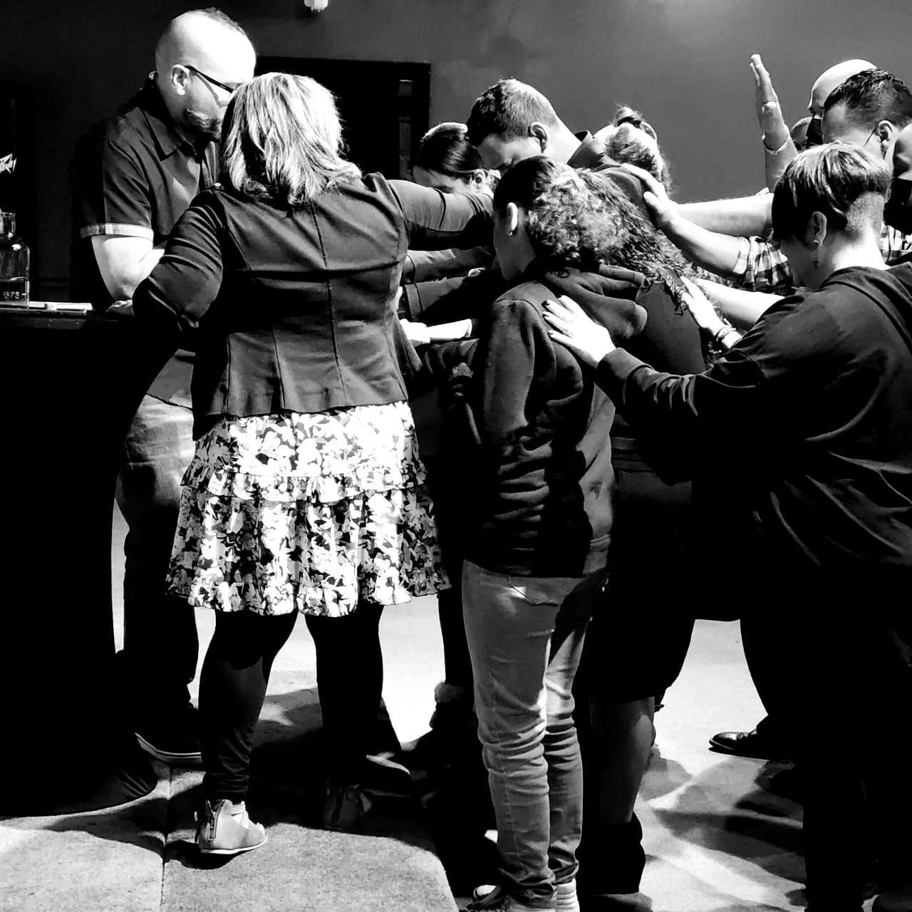 Group of people praying together with hands on each other in a dimly lit room.