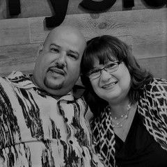 Couple smiling together in a room. Man with a patterned shirt, woman with glasses and pearls.