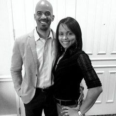 Smiling couple posing. Man in blazer and shirt, woman in black top. White paneled wall backdrop.