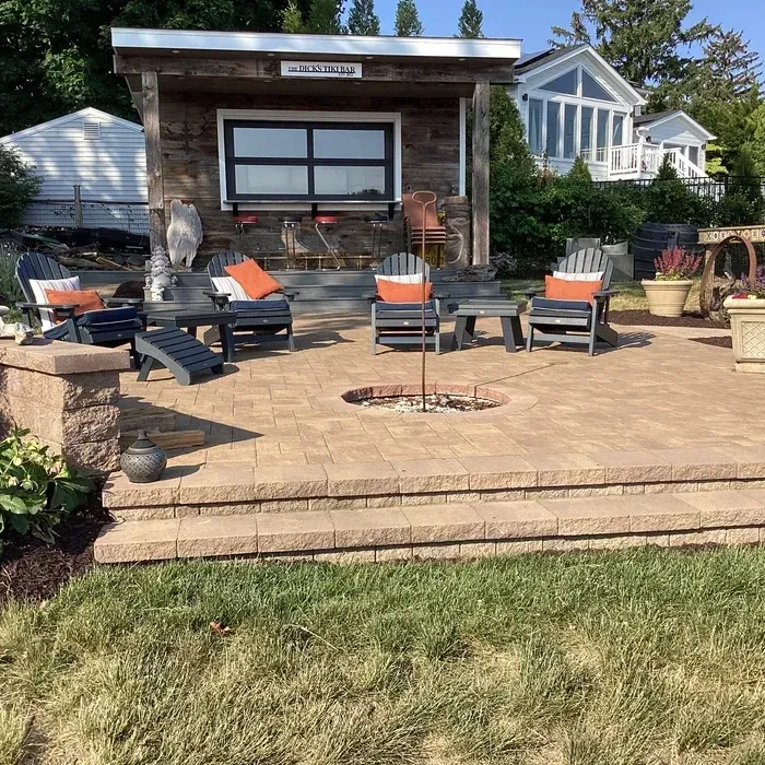 A patio with a fire pit, several outdoor chairs, and a rustic wooden cabana, set on a grassy lawn with a house behind.