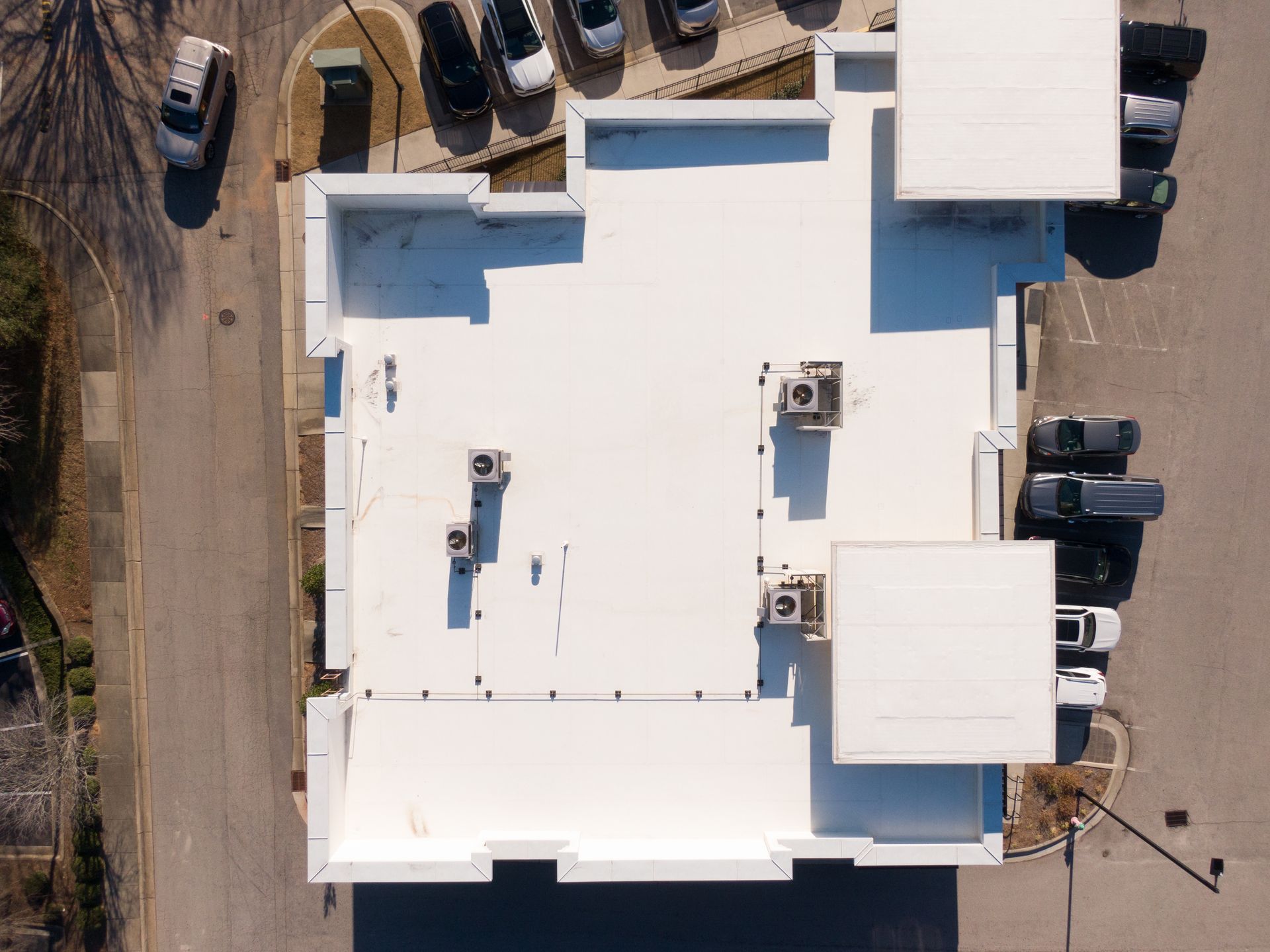 Vue aérienne du toit d'un immeuble commercial blanc avec unités de climatisation et parking environnant avec voitures.
