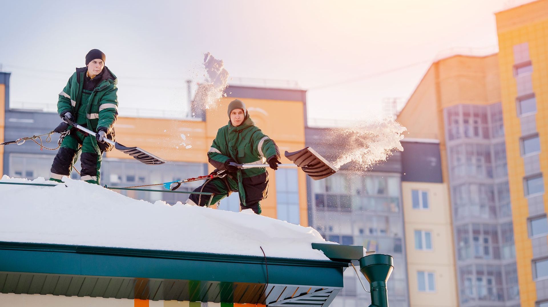 Two workers in green coveralls shoveling snow off a rooftop in front of a building; snow is flying.