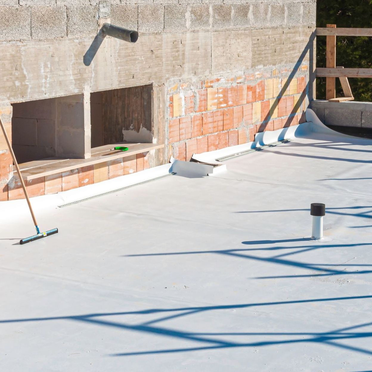 Toit plat en construction avec revêtement gris, entouré de murs en briques et en béton. Ombres projetées par les outils.