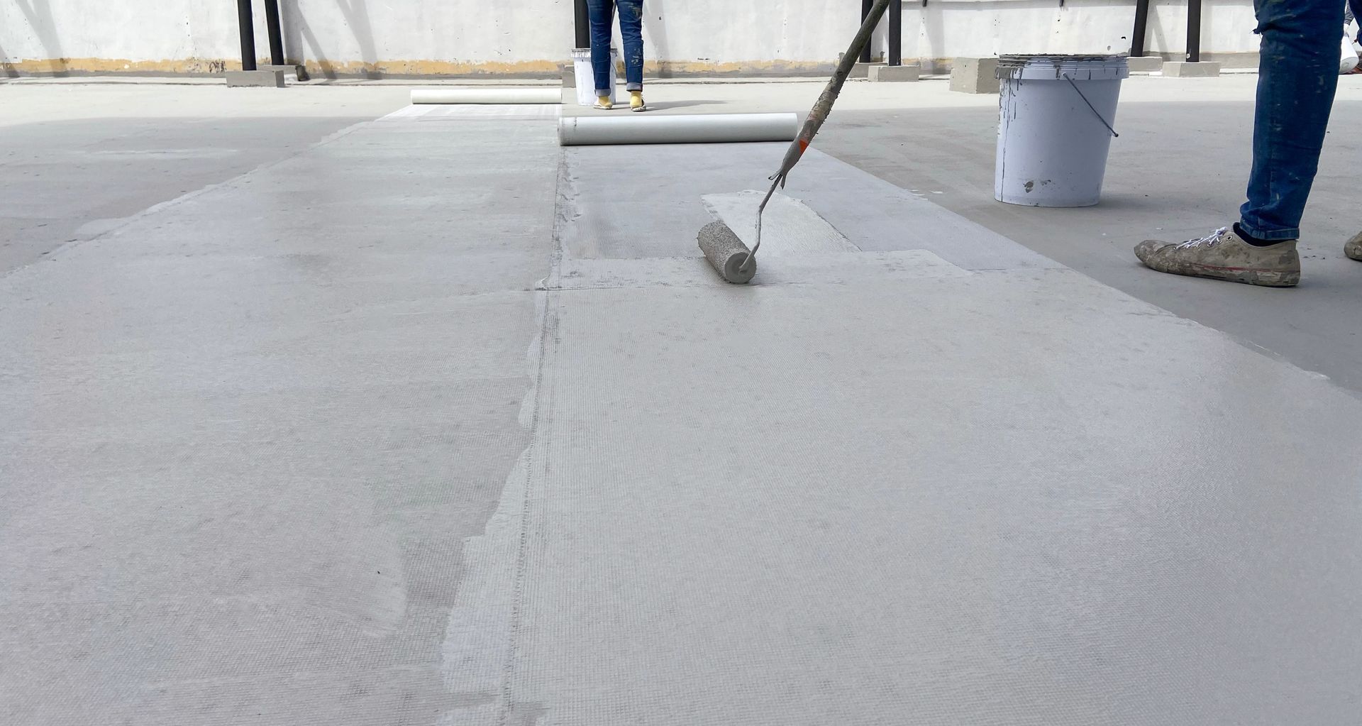 Une personne applique du mastic blanc sur une surface en béton gris, vraisemblablement un toit, avec un seau à proximité.