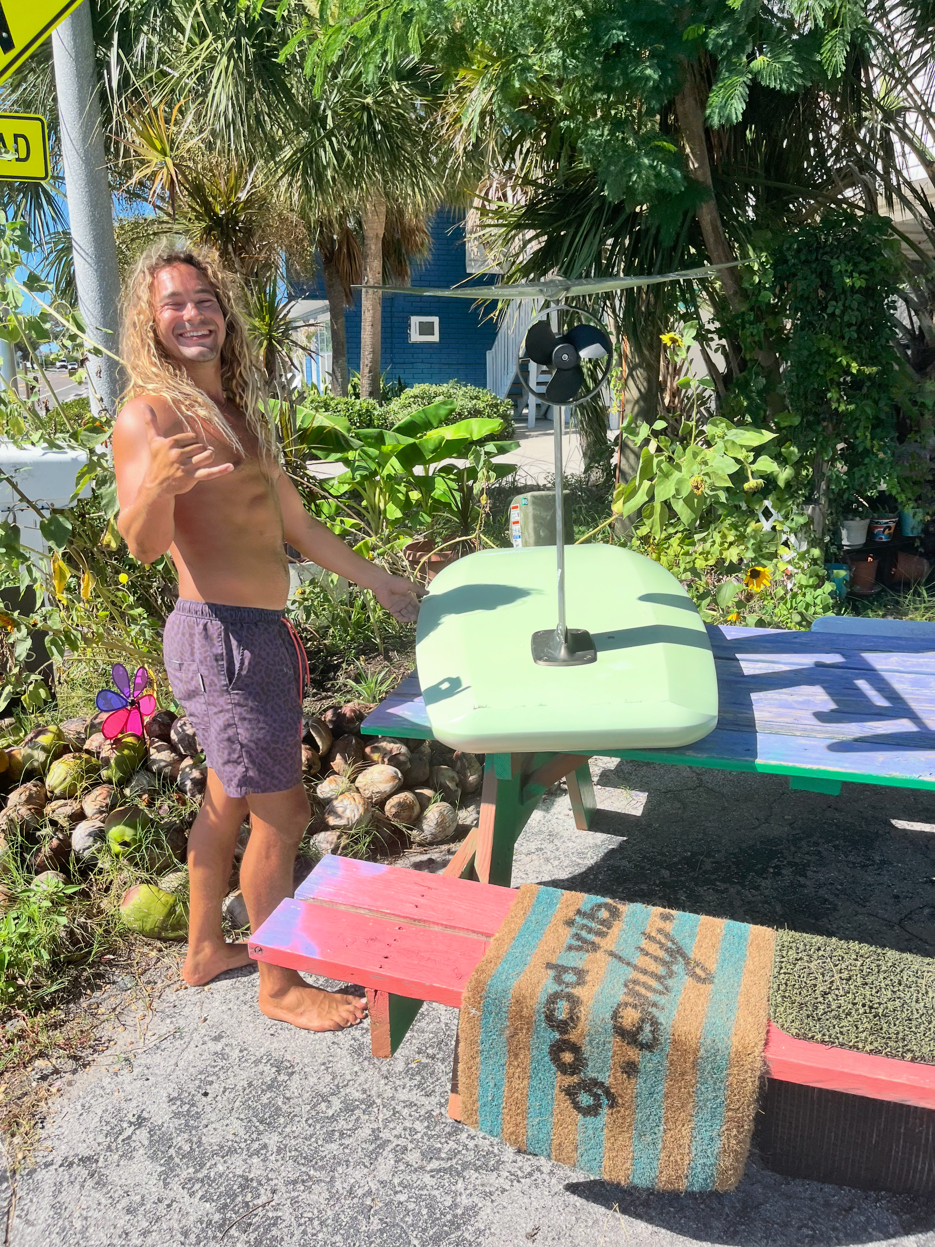 Man with long hair smiles, posing shirtless next to colorful picnic table and bench with hydrofoil