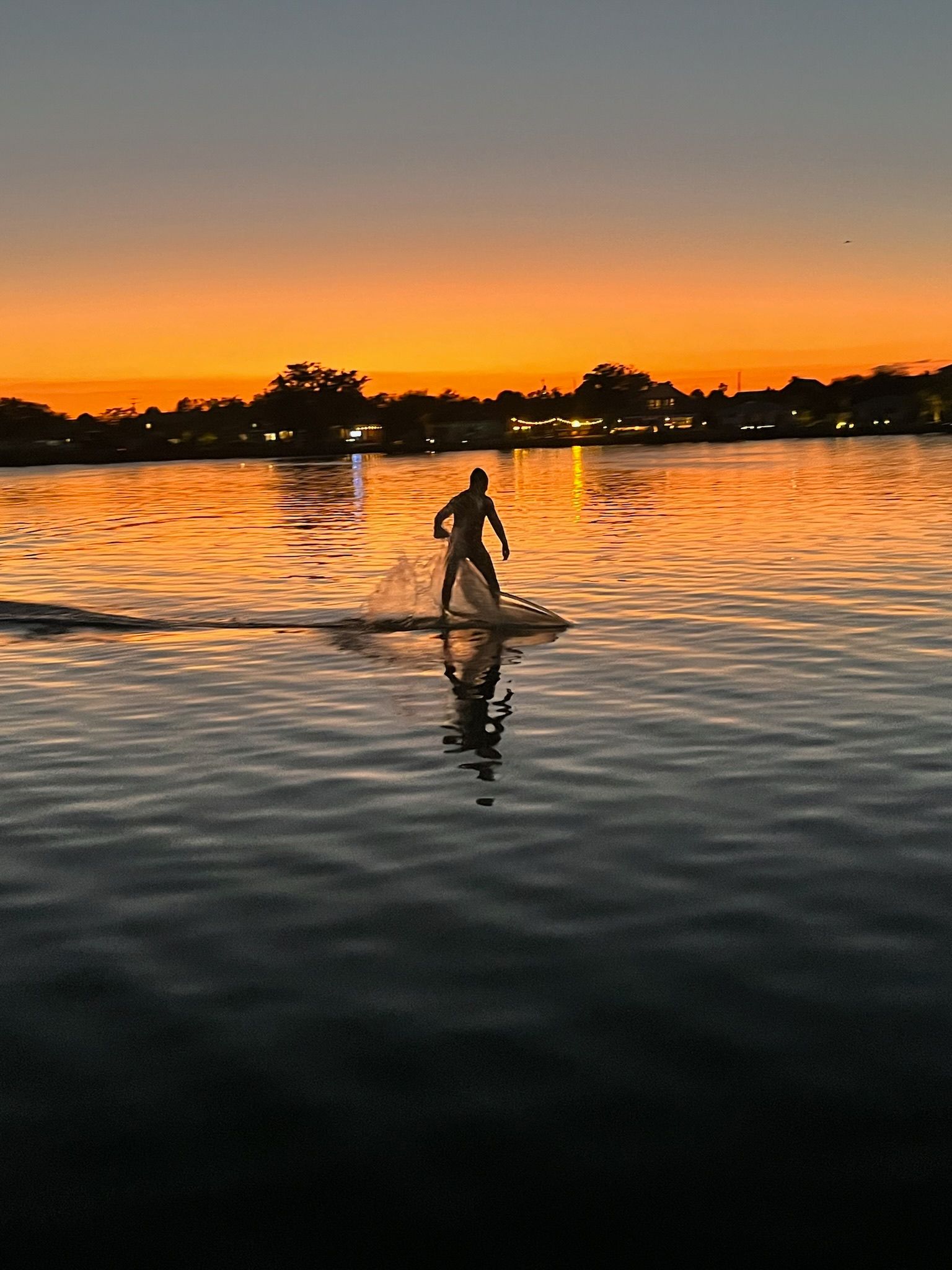 Silhouette of person on hydrofoil electric surf boad at sunset, with orange sky reflecting on water.