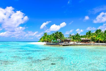 Turquoise ocean meets white sand beach under a bright blue sky with fluffy clouds; tropical island resort.