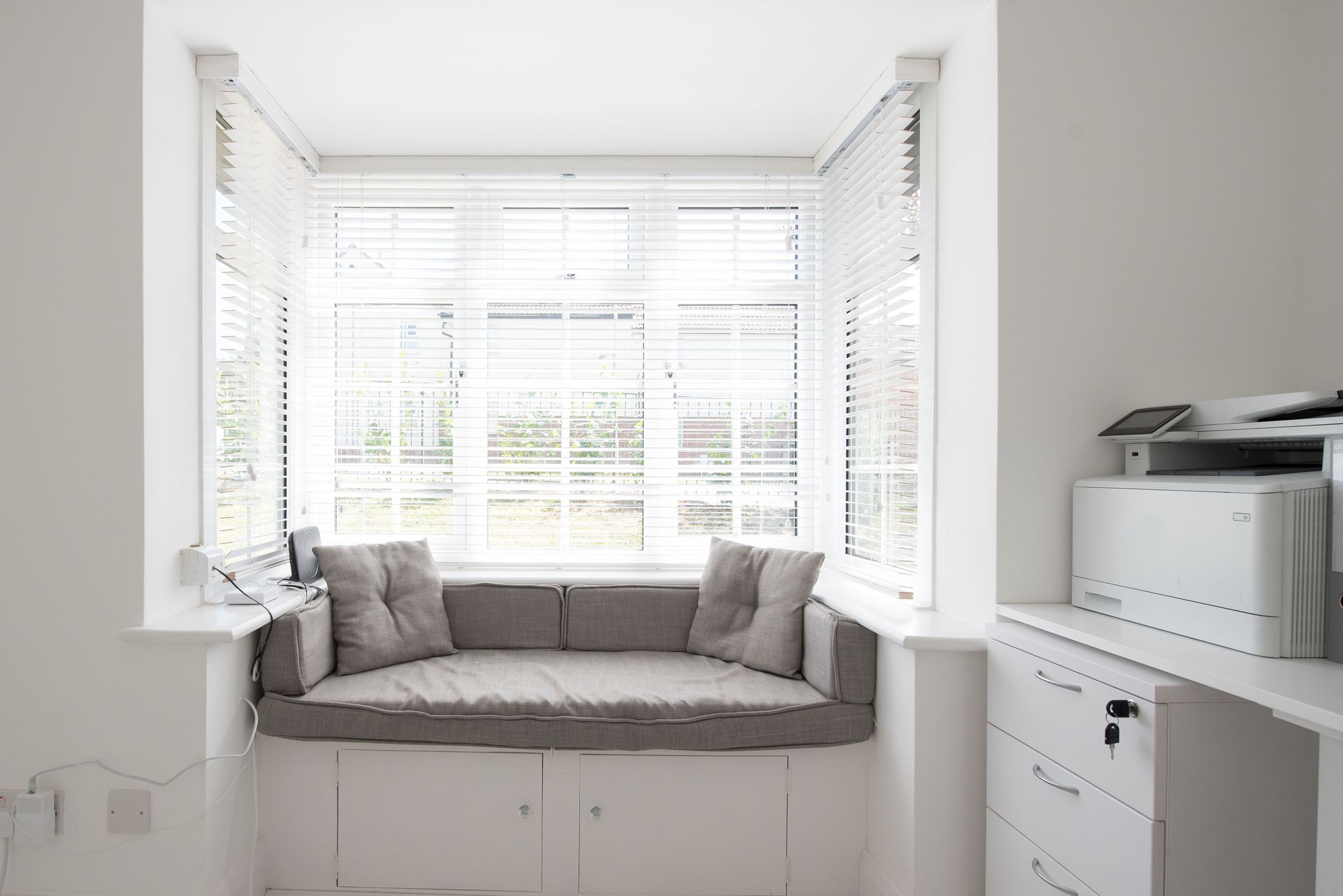 Bright window seat with grey cushions and white blinds.  White desk with a printer to the right.