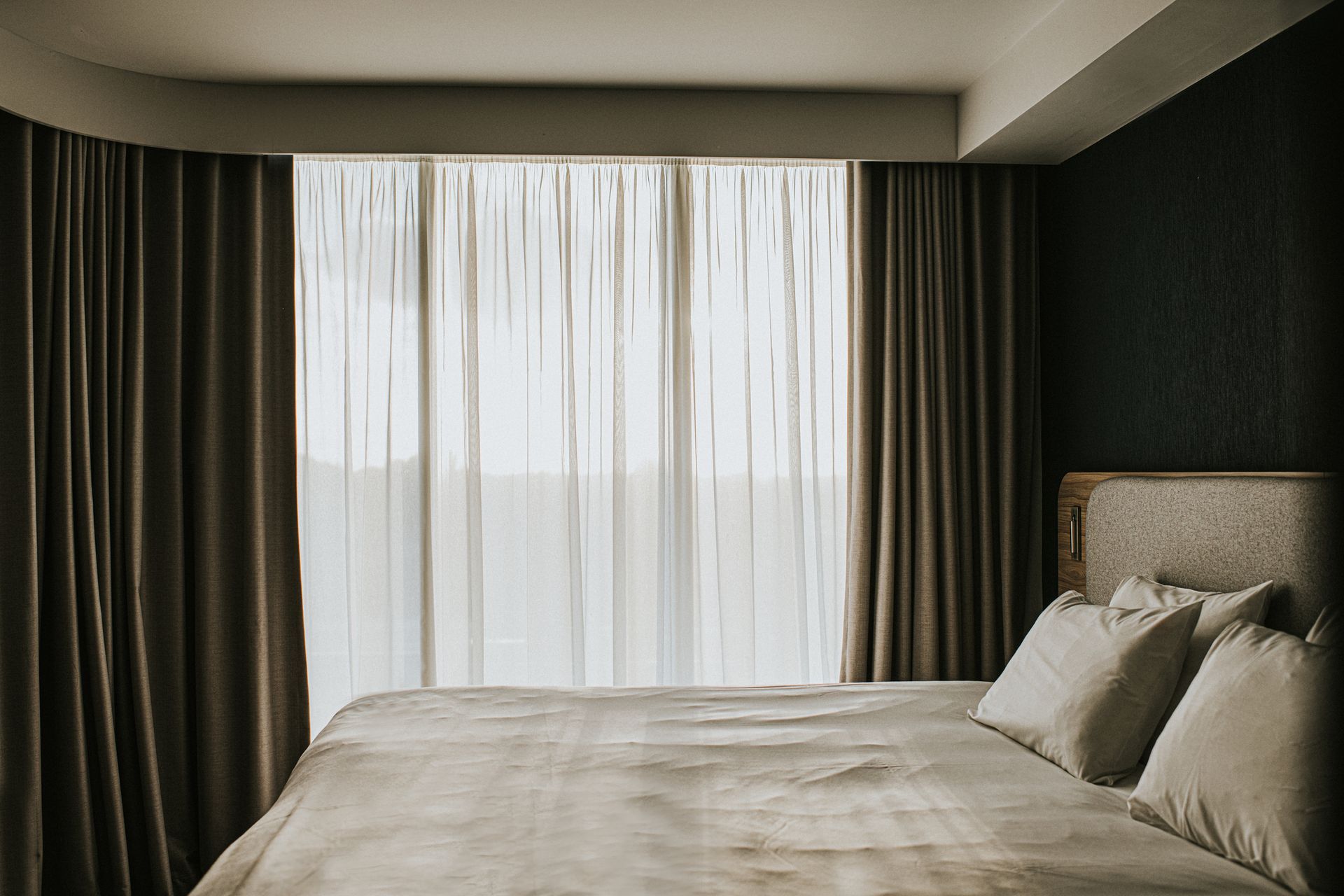 Bedroom with bed, neutral bedding, sheer curtains, and dark drapes framing a window.