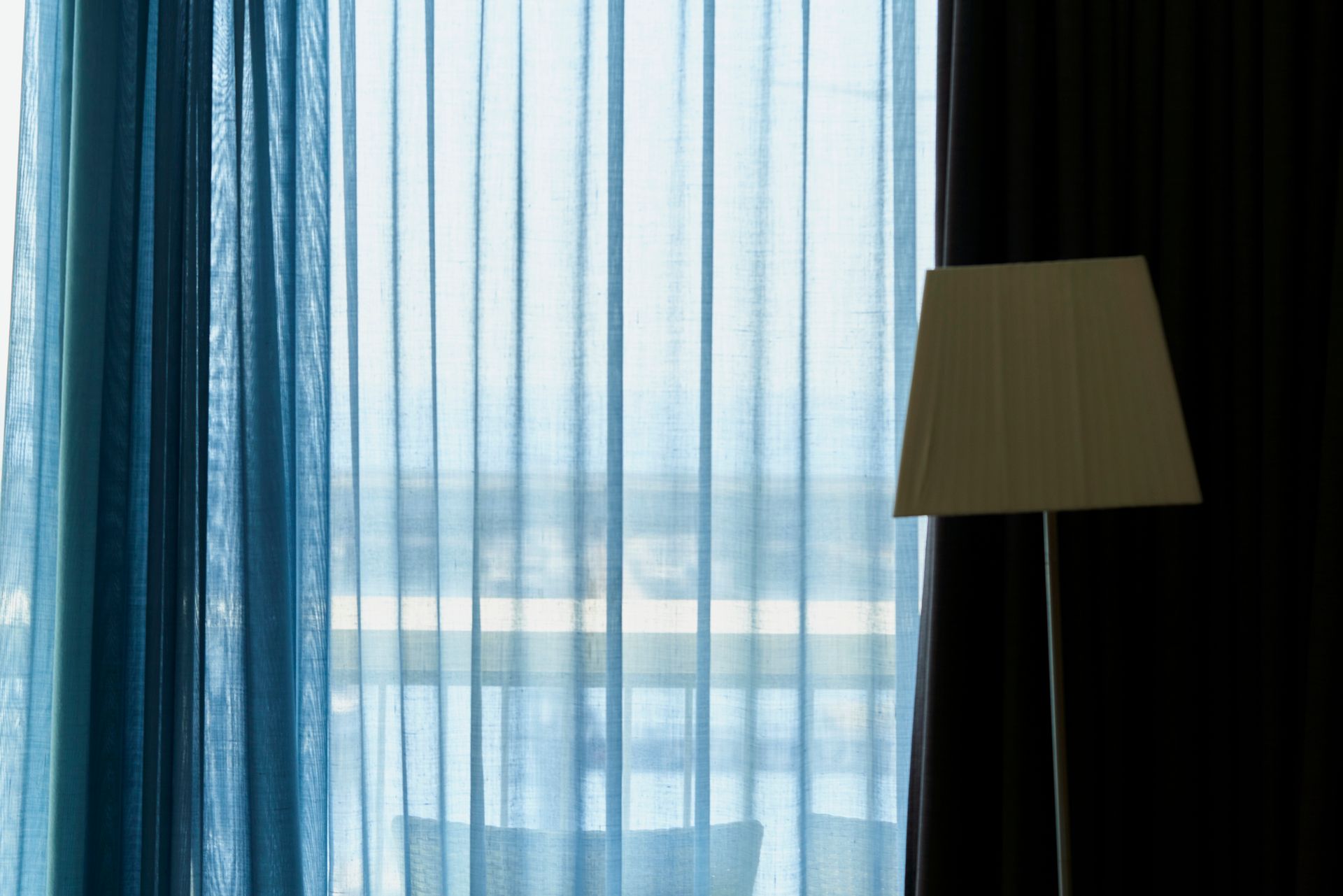 Blue sheer curtains in front of a window, with a floor lamp to the right against a dark curtain.