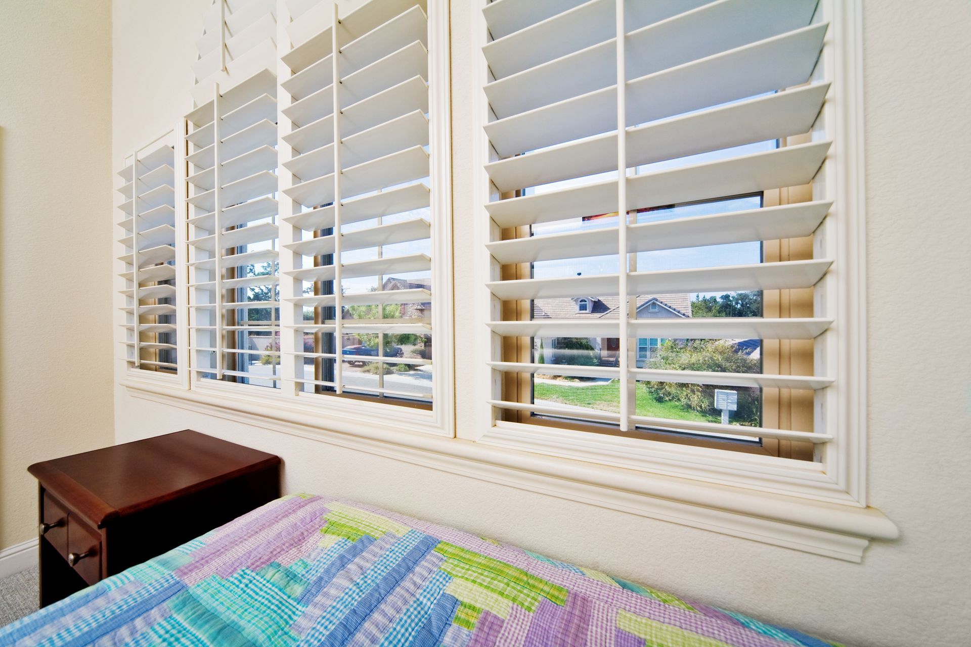 Bedroom window with white shutters; small nightstand; multi-colored quilt on the bed.