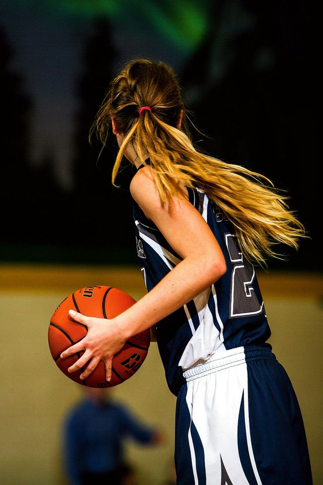 Basketball player in dark jersey dribbling on the court, seen from the side with long hair flowing behind.