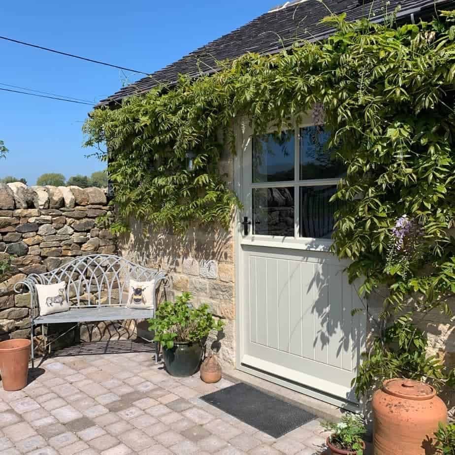 Stone cottage entrance with climbing greenery, brick patio, bench, and potted plants.