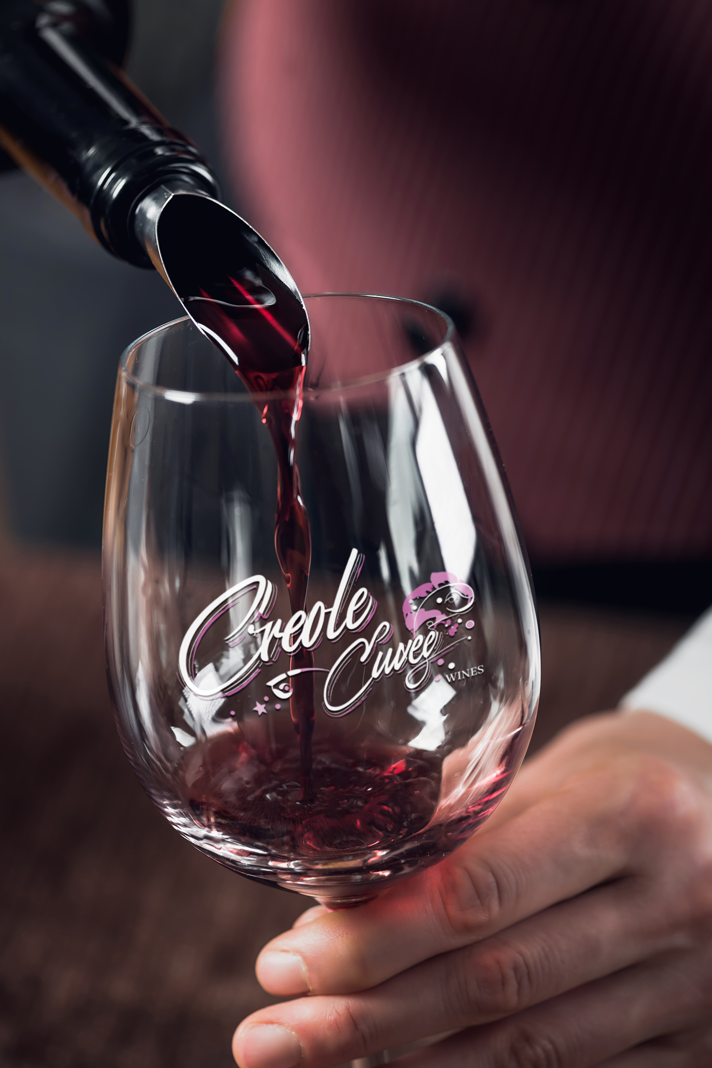 Red wine being poured into a branded wine glass, held by a person in a burgundy top.