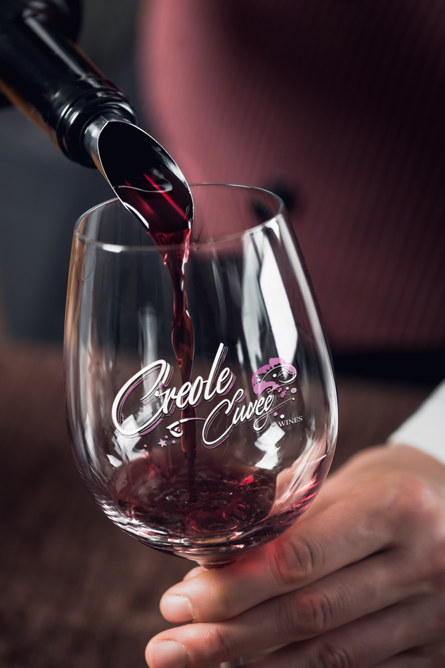 Red wine being poured into a branded wine glass, held by a person in a burgundy top.