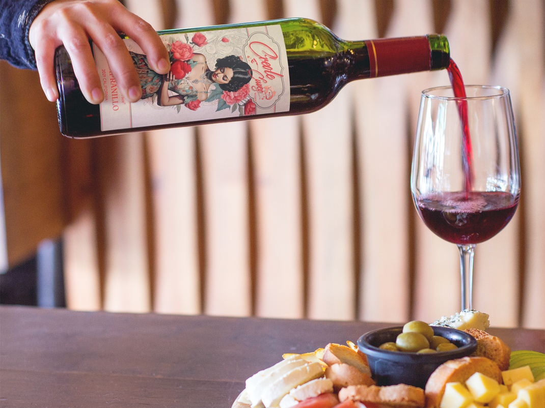 A person pouring red wine into a glass next to a charcuterie board with olives and cheese.