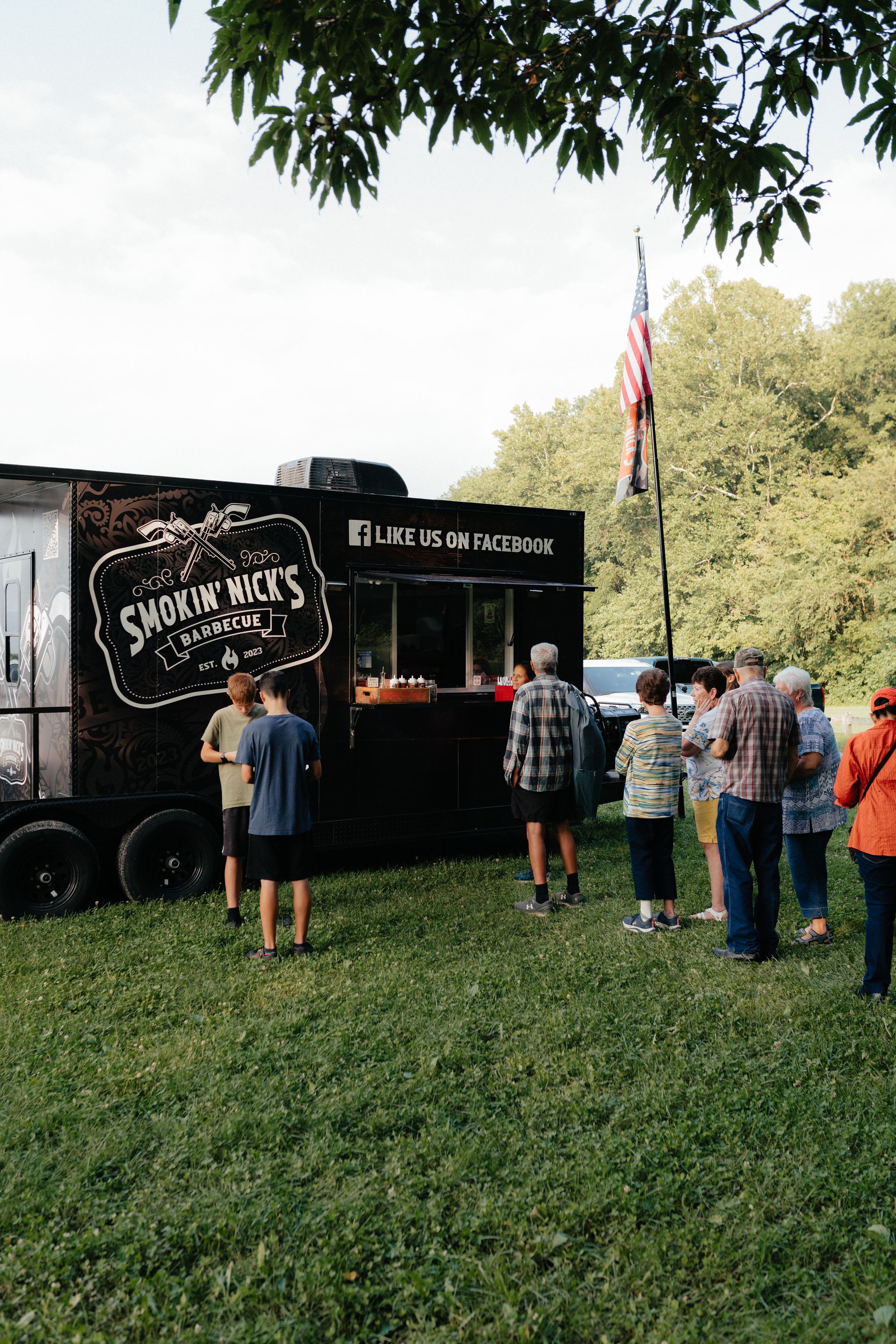 People line up at a food truck in a grassy park, an American flag flies overhead.