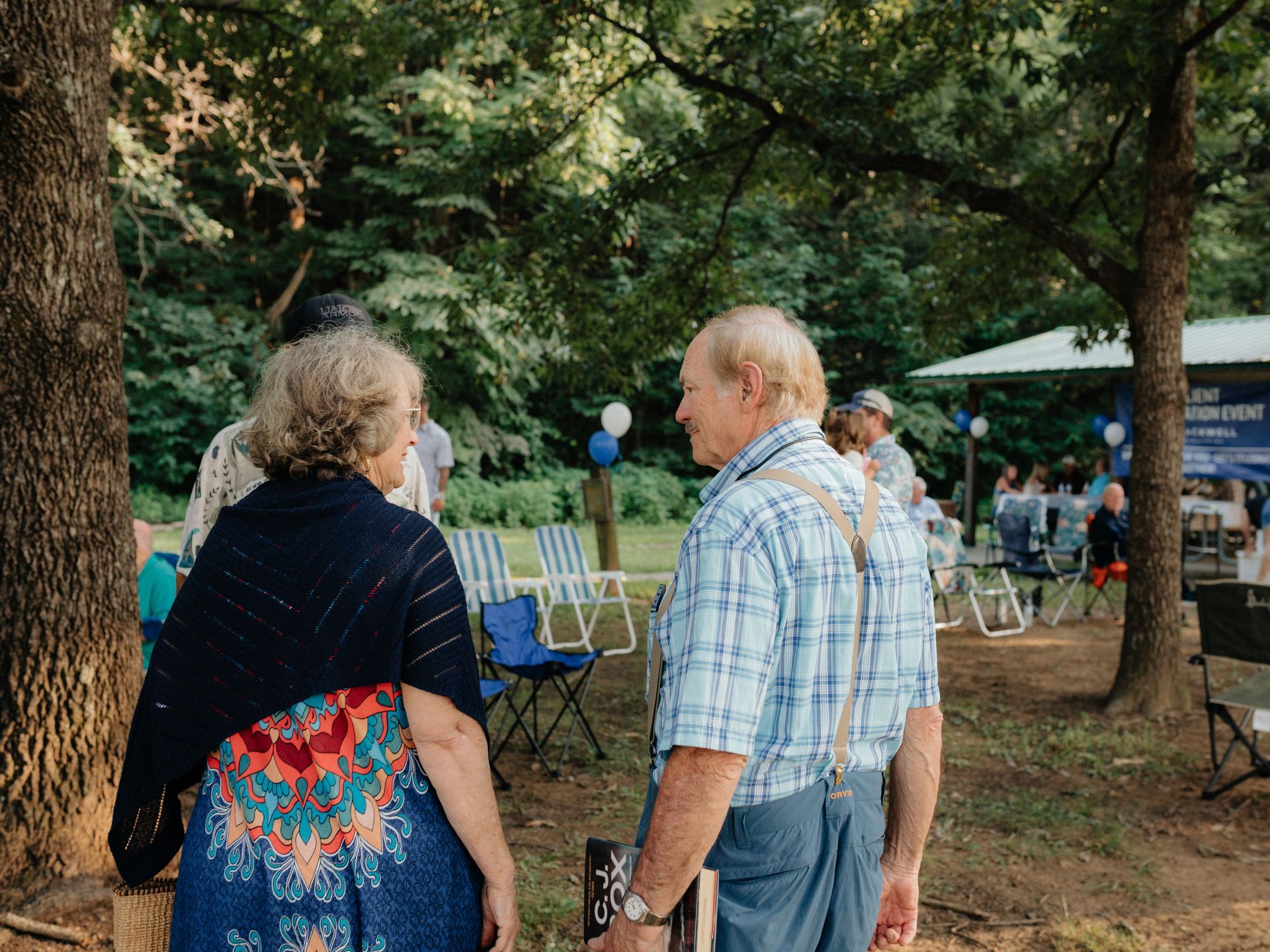 Elderly couple conversing at an outdoor event; blue and white color scheme, trees, lawn chairs, and people in the background.