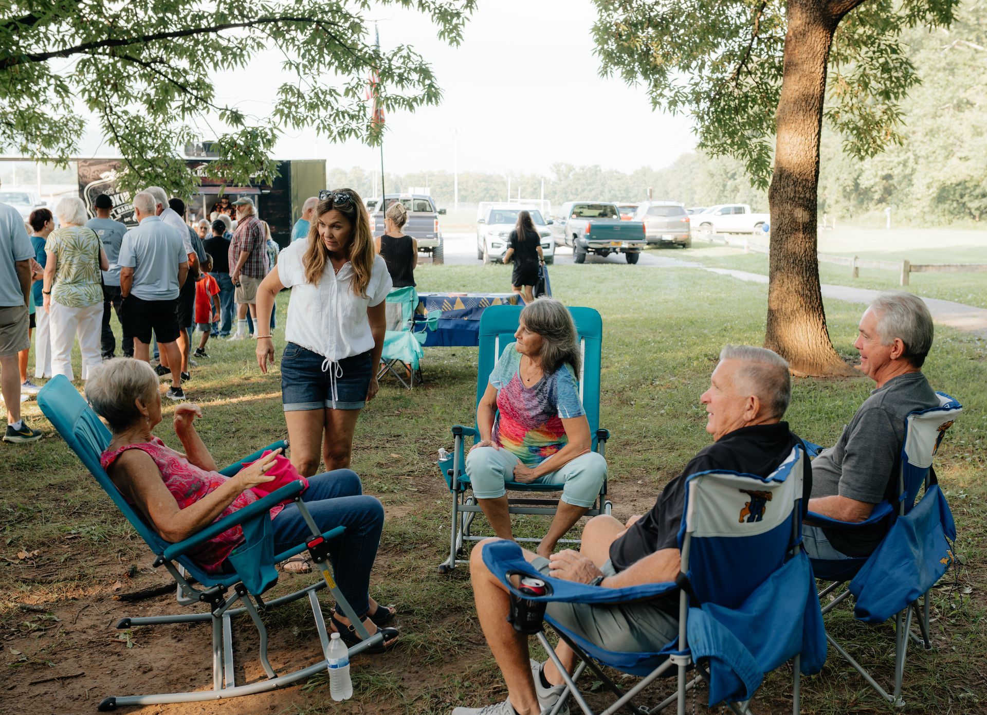 People relaxing in lawn chairs at an outdoor gathering. Some are talking and smiling. Trees and cars are in the background.