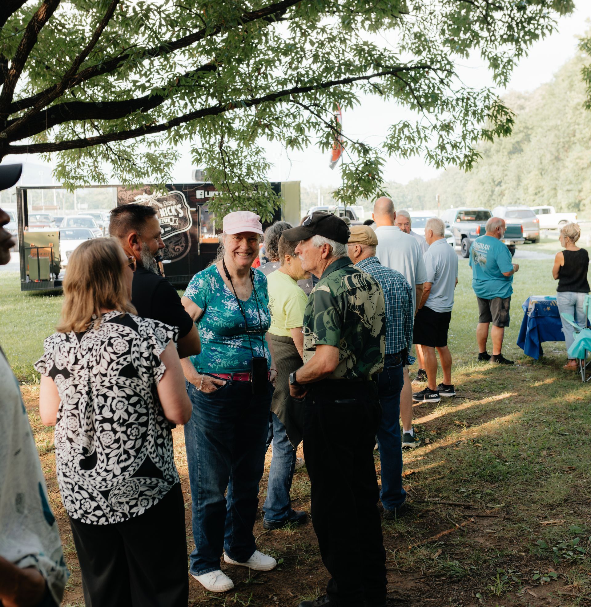 Group of people socializing outdoors near a trailer; trees and greenery in background.