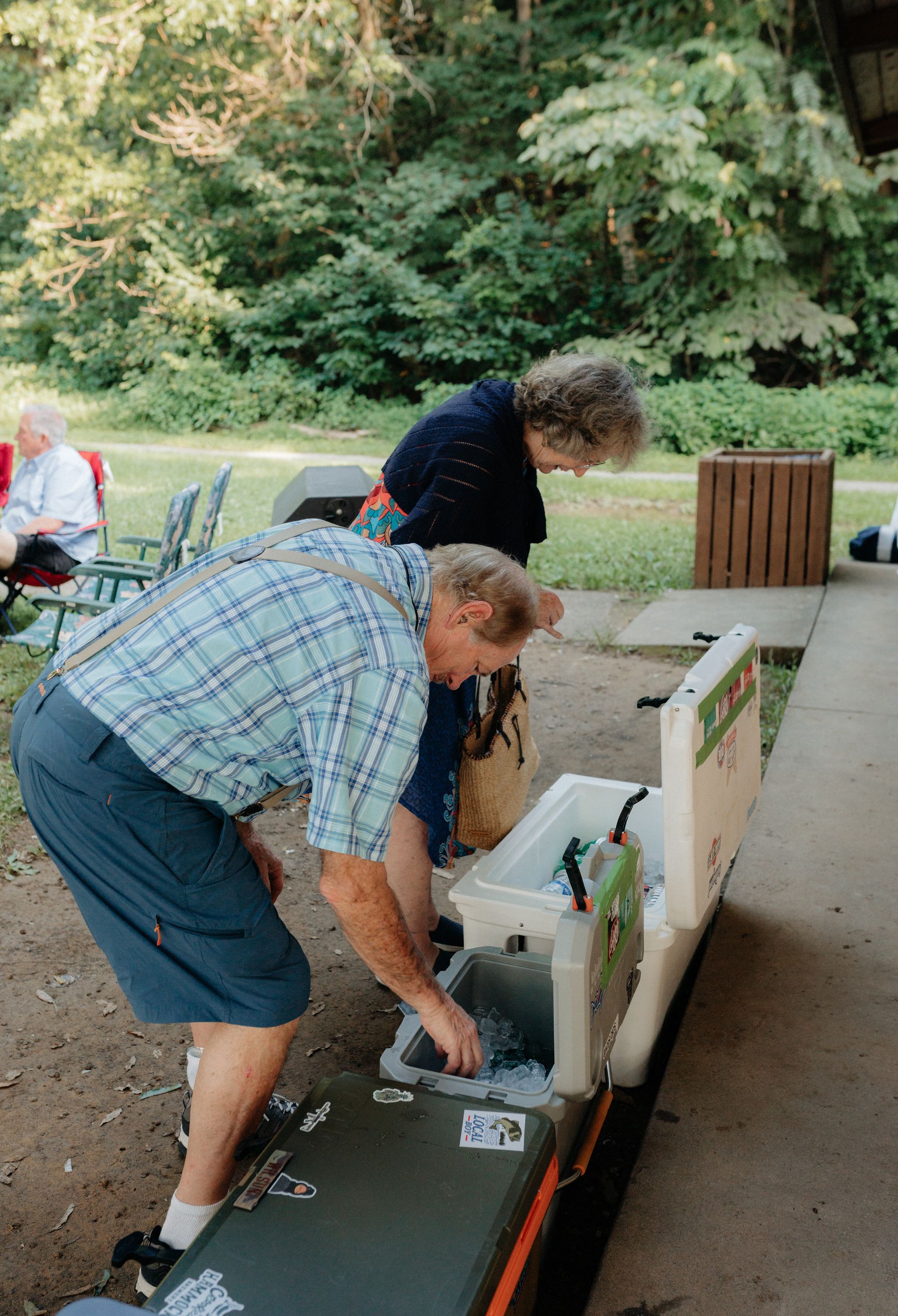Man and woman at picnic table, selecting from a cooler. Outdoors, trees in background.