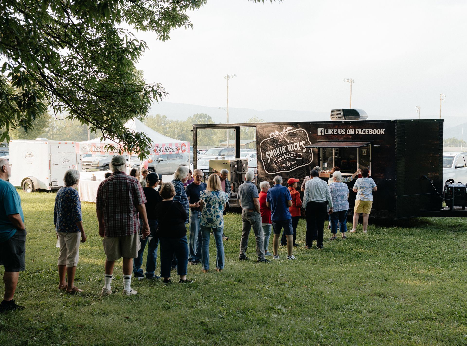 People line up at a black food truck in a park, sunny day.